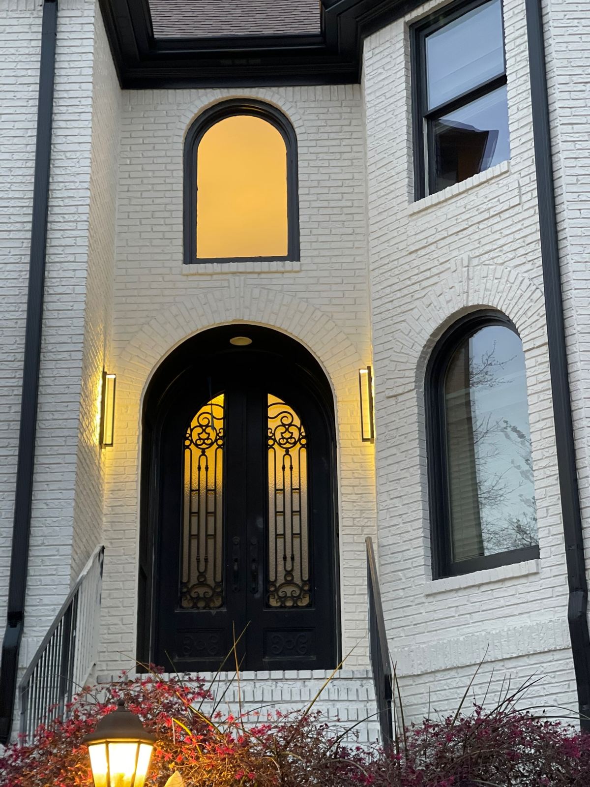 White brick home entry with arched iron double doors and lit sconces
